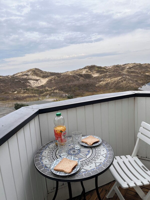 A balcony with a table set for two, a bottle of juice, and a plate of toast. The view includes a sandy landscape and a cloudy sky.