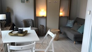 A modern living room with a white dining table, a grey sofa, and a white chair. There are wall-mounted shelves with books and decorative items.