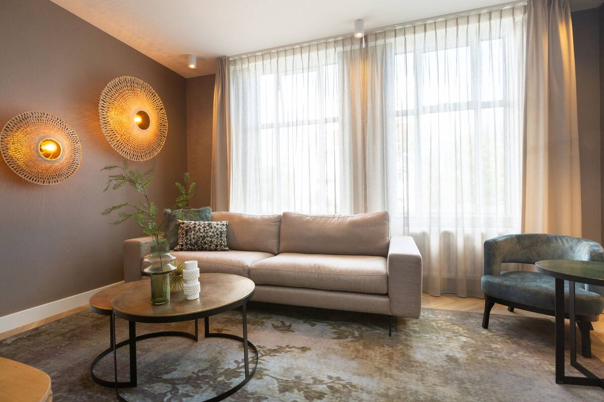 A living room with a sofa, a coffee table, and a chair. There are two wall sconces with lights, a window with sheer curtains, and a plant on the coffee table.