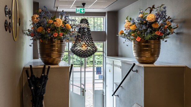 Un pasillo con dos grandes arreglos florales sobre pedestales, una lámpara colgante y una puerta que da al exterior.