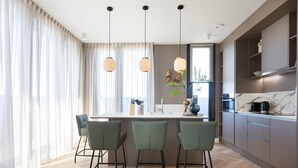 Cocina moderna con isla central, taburetes de bar, luces colgantes y vistas al exterior.