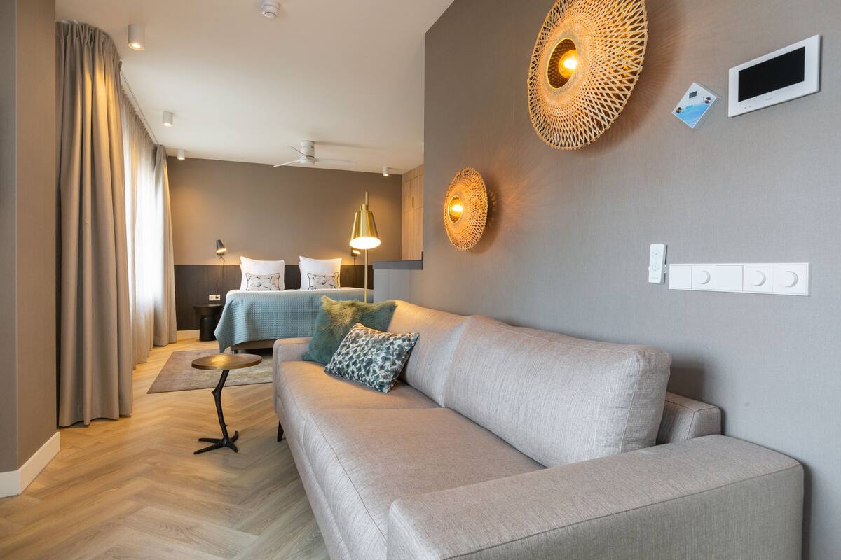 A modern hotel room with a sofa, a small round table, a bed with a blue bedspread, and a wall-mounted thermostat.