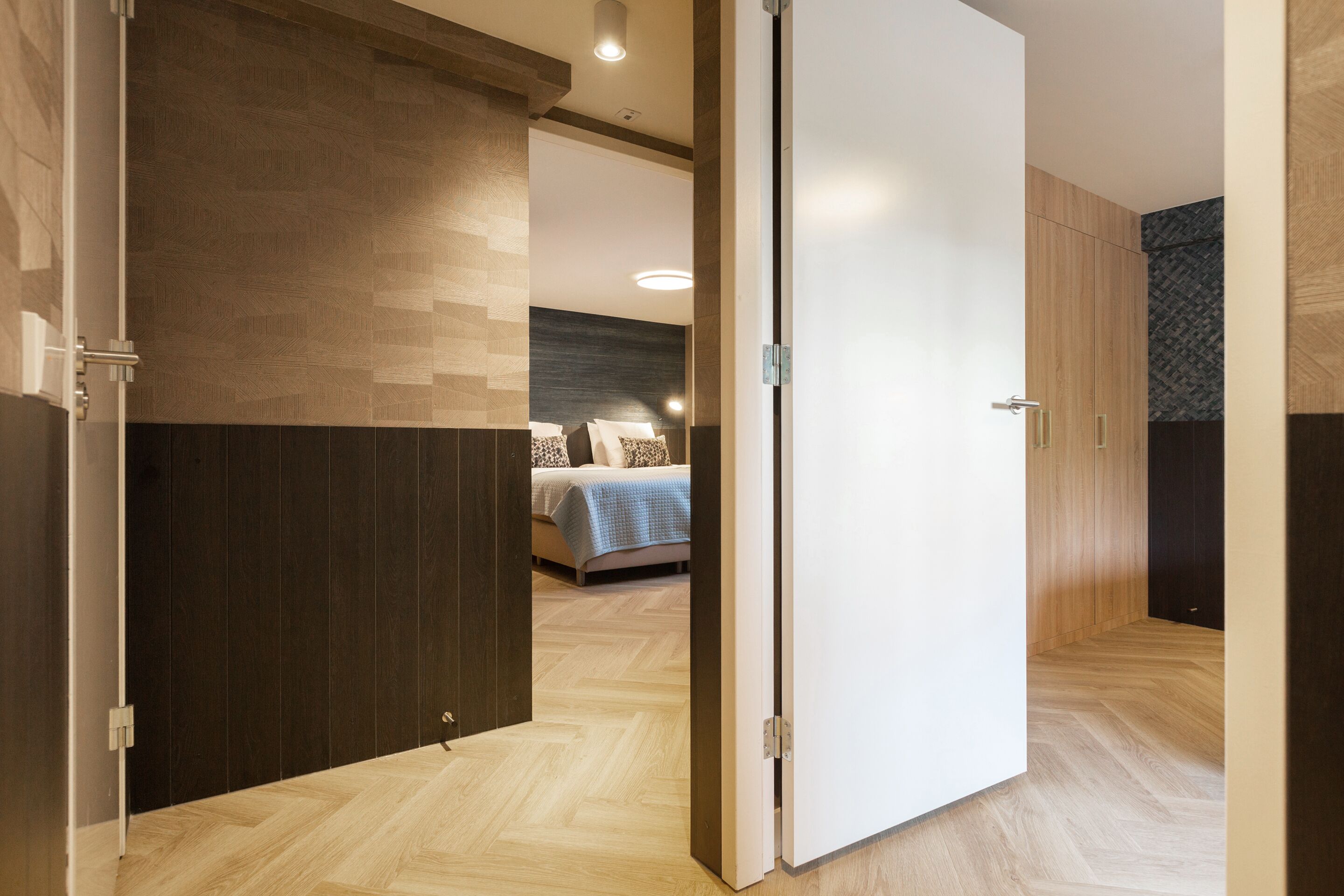 A modern hotel room with a wooden floor, a bed with a blue bedspread, and a dark wall with a geometric pattern.