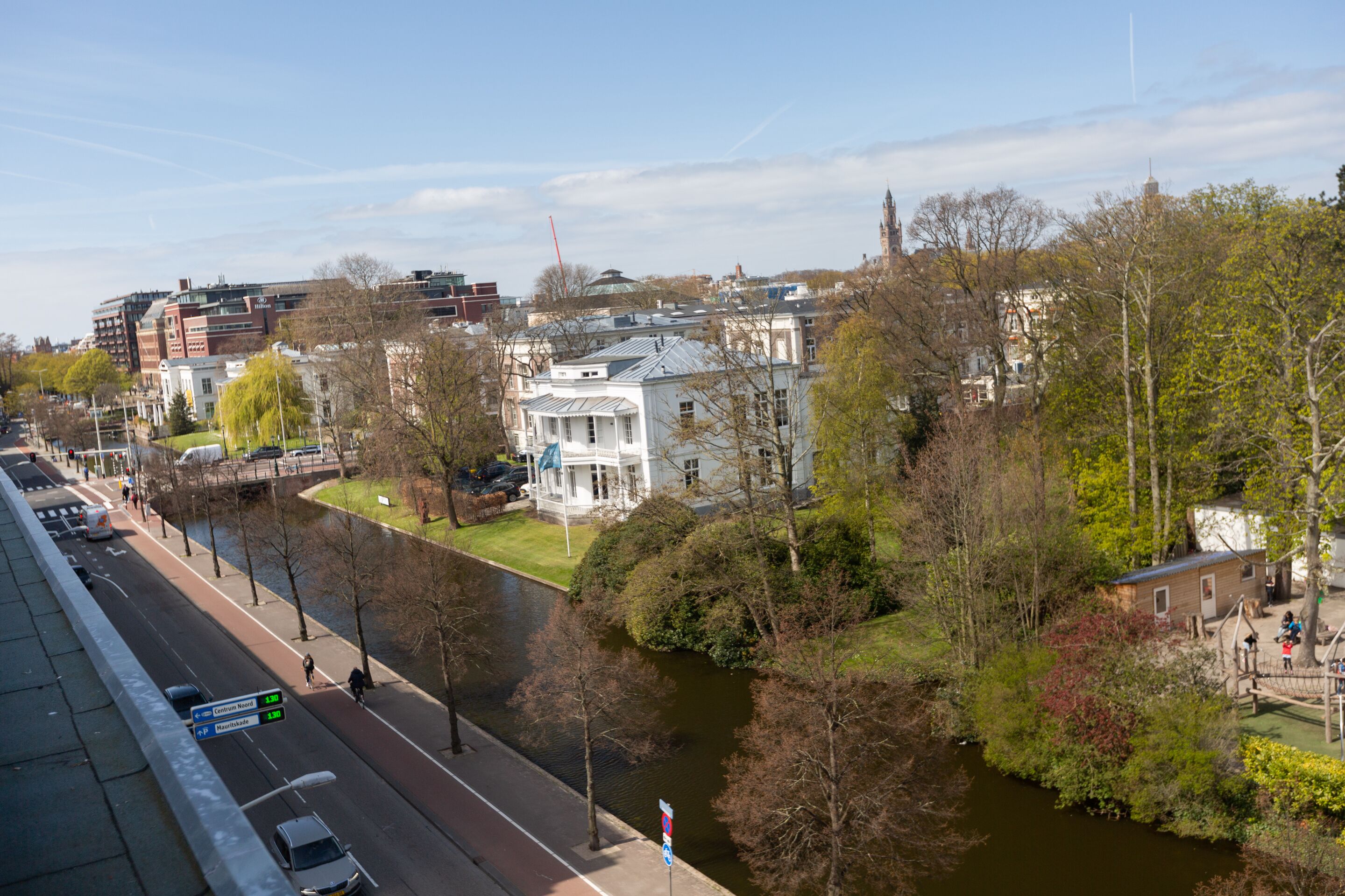 A riverside view of a historic building with a large lawn, a bridge, and a modern building in the background.