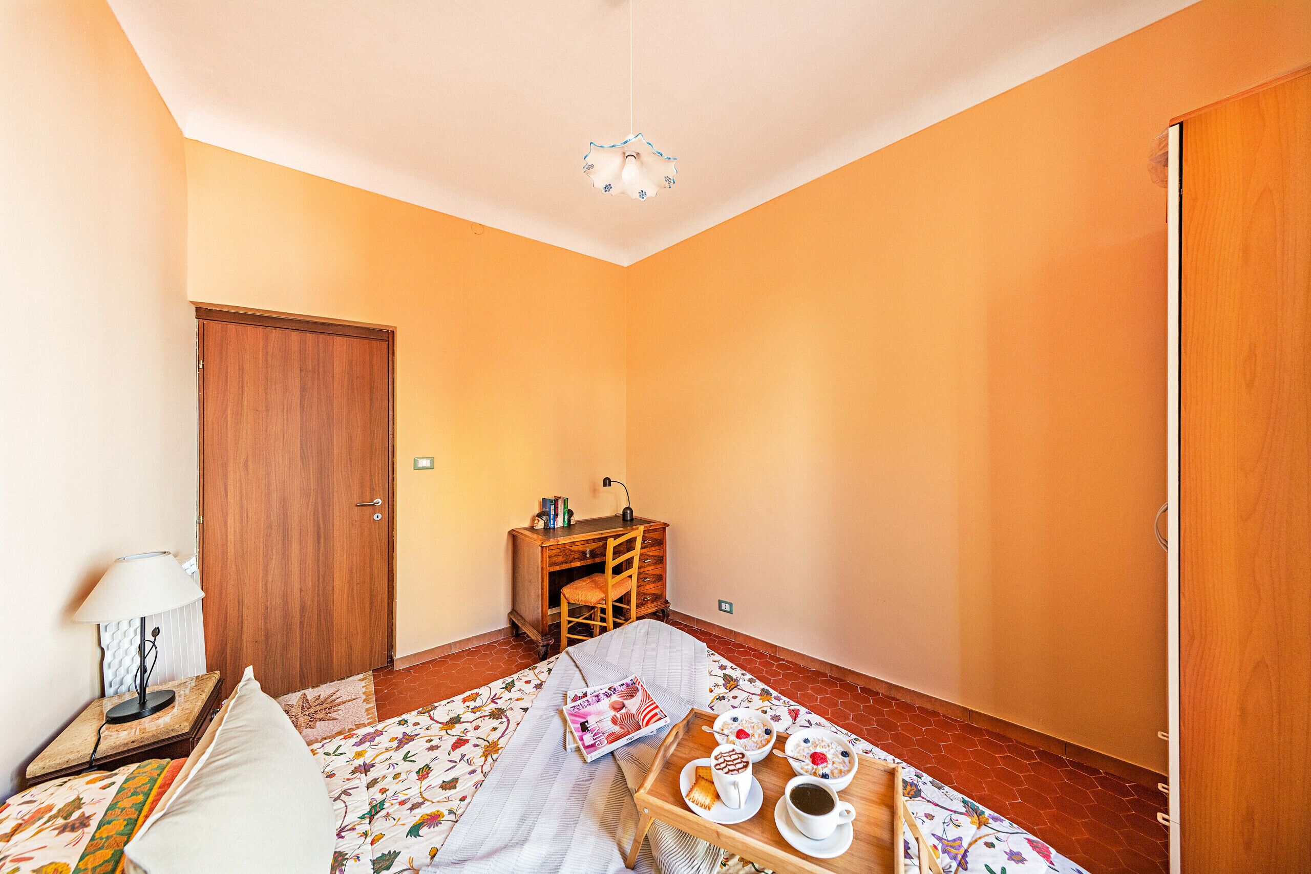 A room with a bed, a wooden desk, a chair, a table with a tray of breakfast items, and a hanging light fixture.