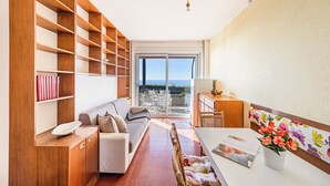 Un salon moderne avec un canapé, des étagères en bois, une table à manger et une vue dégagée sur l’extérieur grâce à une grande porte vitrée.