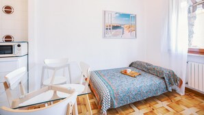 A small, bright room with a bed, a chair, a table, and a picture on the wall.