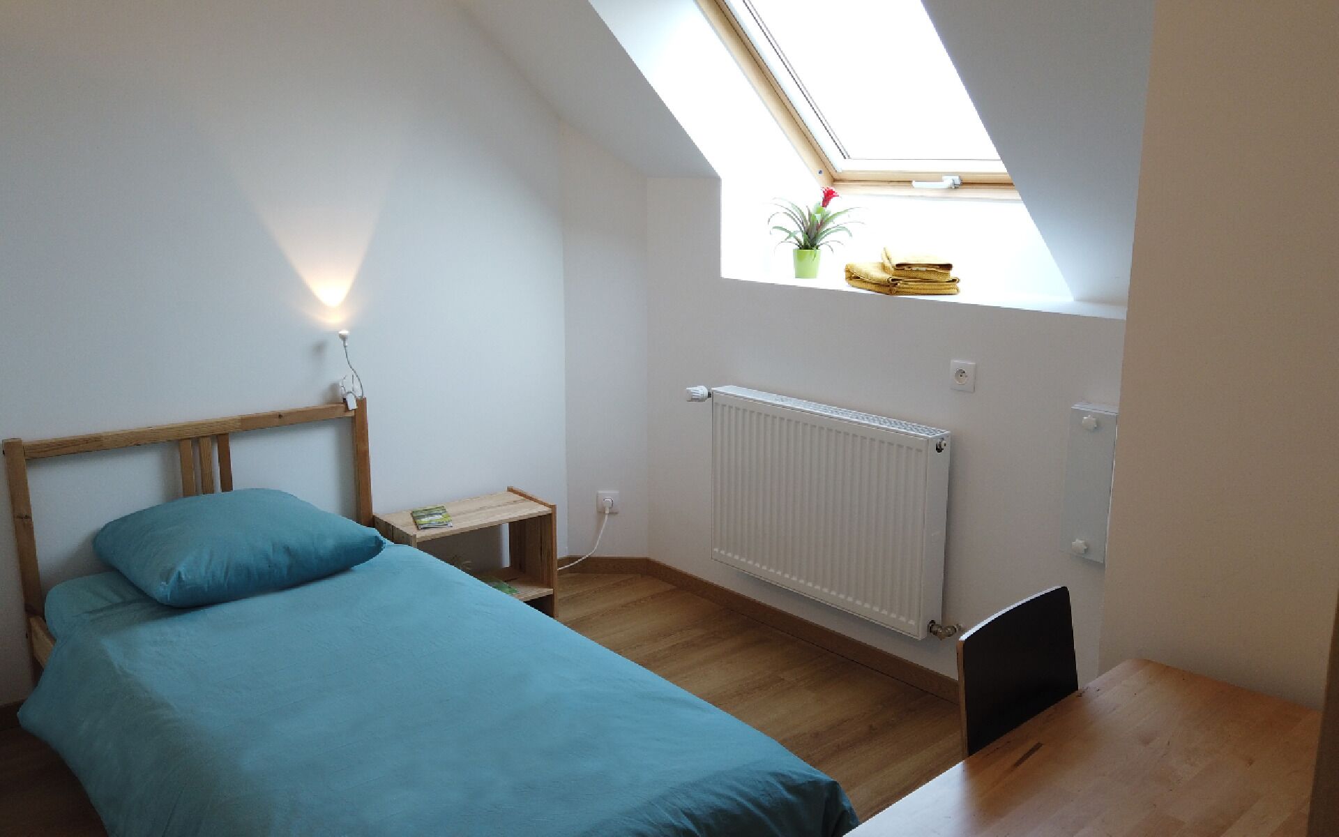 Ein Schlafzimmer mit Dachfenster, einem Bett, einem Nachttisch, einem Heizkörper und einem kleinen Fenster mit Pflanze.