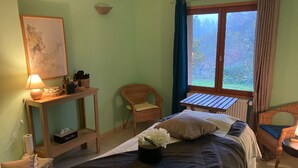 A room with a bed, a small table with a lamp, a wicker chair, and a window with curtains. - AN OASIS WHERE TO FIND THE ESSENTIALS<br>AND TAKE A BREAK IN CONSCIOUSNESS & IN (SAINT USUGE)