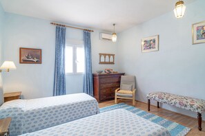 Ein Schlafzimmer mit zwei Betten, einem Schminktisch, einem Stuhl, einer Bank und einem Fenster mit Vorhängen.
