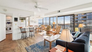 Een moderne woonkamer met een bank, een eethoek met stoelen, een keuken met een koelkast, en grote ramen met uitzicht.