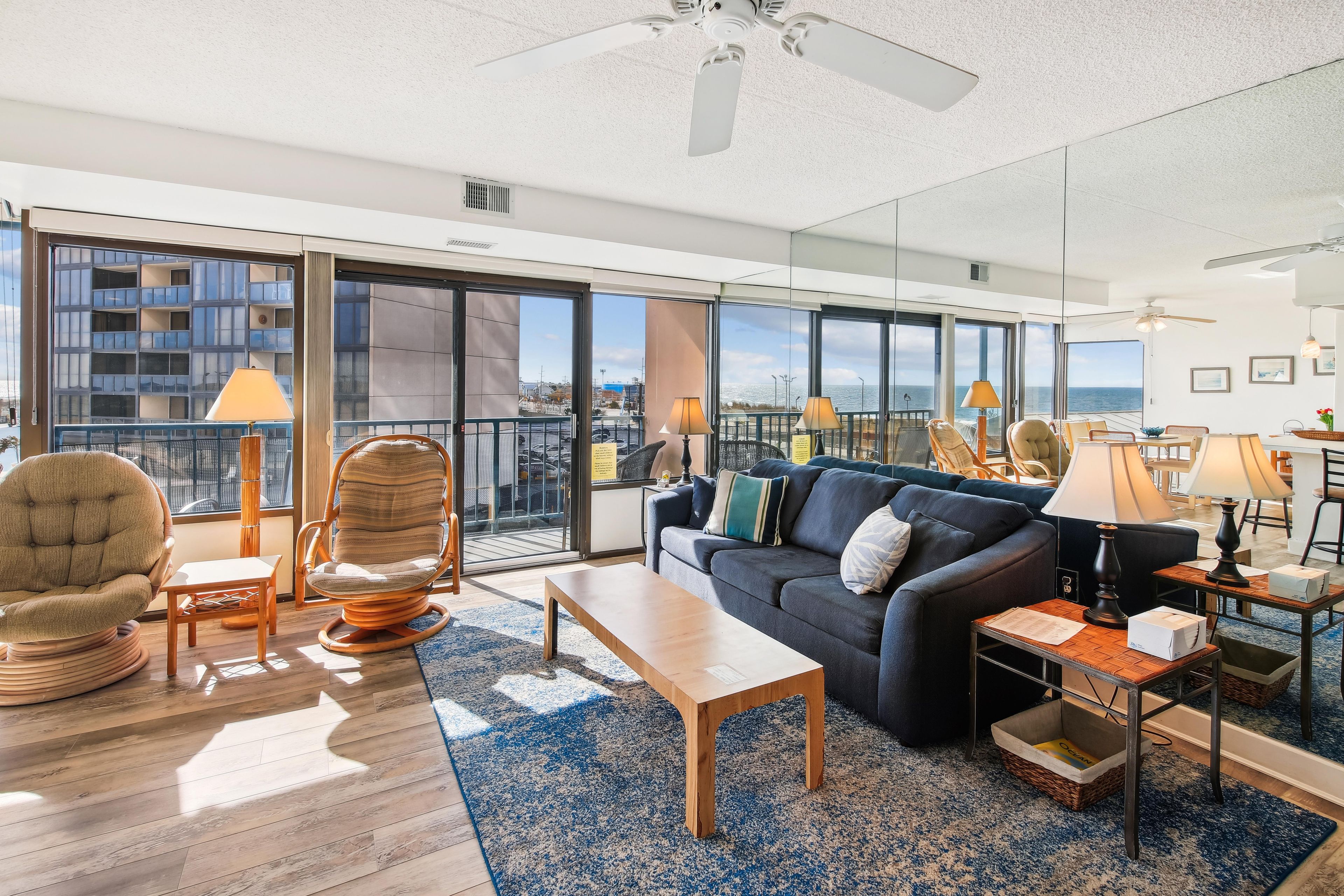 A spacious living area with a large sofa, a coffee table, and a dining table. There are two chairs, a floor lamp, and a ceiling fan. The room has large windows offering a view of the city and the sea.