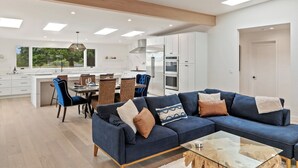 A modern living room with a blue sofa, dining area, kitchen, and large windows. - Pacific Jewel: Brand New Build with Views (Monterey)