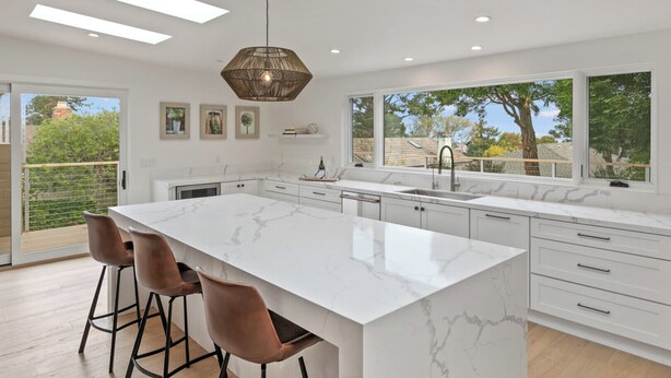 A modern kitchen with a large island, bar stools, a hanging light fixture, and a view of the outdoors. - Pacific Jewel: Brand New Build with Views (Monterey)