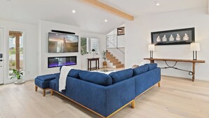 A modern living room with a blue sectional sofa, a fireplace, a TV, and a staircase. - Pacific Jewel: Brand New Build with Views (Monterey)