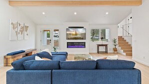 A modern living room with a large blue sofa, a fireplace, a TV, and a staircase. - Pacific Jewel: Brand New Build with Views (Monterey)