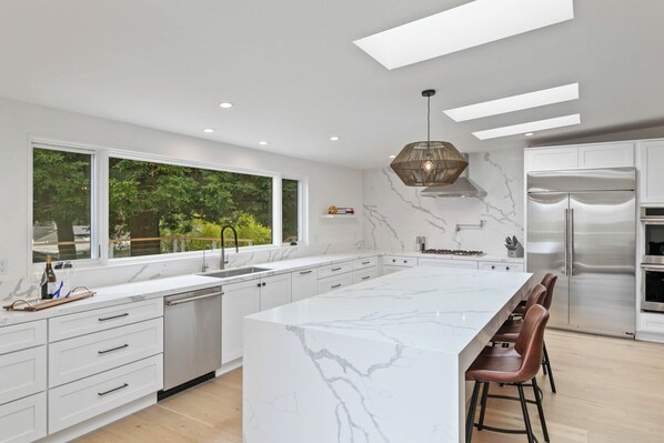 A modern kitchen with a large island, stainless steel appliances, and a view of greenery outside.