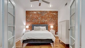 A bedroom with a brick wall, a bed with white bedding, two bedside tables with lamps, and a ceiling fan. - Elegant 2BR condo 3 blocks to French Quarter. King suite, garage parking, walk to Bourbon. (New Orleans)