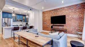 A modern kitchen with blue cabinets, a dining area with a wooden table, and a living room with a brick wall and a flat-screen TV. - Elegant 2BR condo 3 blocks to French Quarter. King suite, garage parking, walk to Bourbon. (New Orleans)