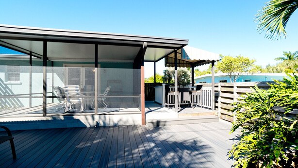A covered outdoor patio with a table and chairs, a wooden deck, and a view of a house in the background.