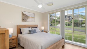 A bedroom with a large window, a bed with a wooden headboard, bedside tables, and a ceiling fan. - Sea Renity - Coastal Hideaway (Bonny Hills)