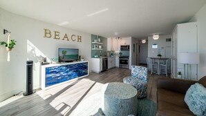 A spacious living area with a kitchen, a dining area, and a large TV screen. - Beach Perfect Top of the Gulf 625-Beautiful Modern Beachfront Studio-Perfect Place in Paradise! (Panama City Beach)