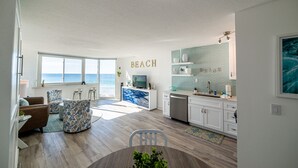 A modern living room with a large window overlooking the ocean, a kitchen with white cabinets and a blue backsplash, and a dining area with a round table and chairs. - Beach Perfect Top of the Gulf 625-Beautiful Modern Beachfront Studio-Perfect Place in Paradise! (Panama City Beach)