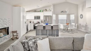 Eine moderne Küche mit weißen Schränken, einer zentralen Kochinsel mit Barhocker, einem Essbereich mit Tisch und Stühlen sowie einem gemütlichen Wohnzimmer mit grauem Sofa und Kamin.