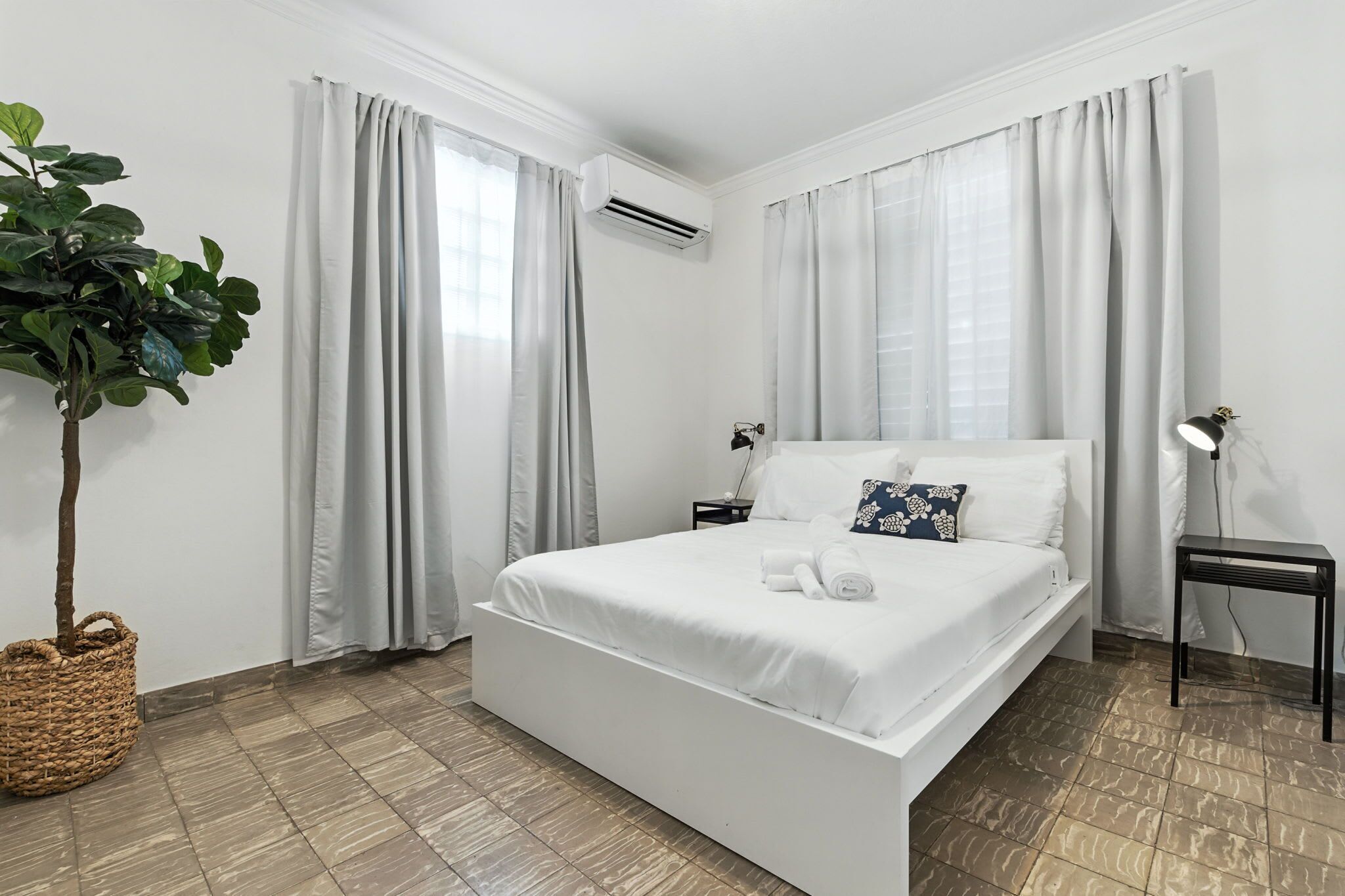 A bedroom with a bed, curtains, an air conditioner, a nightstand, a lamp, and a plant.