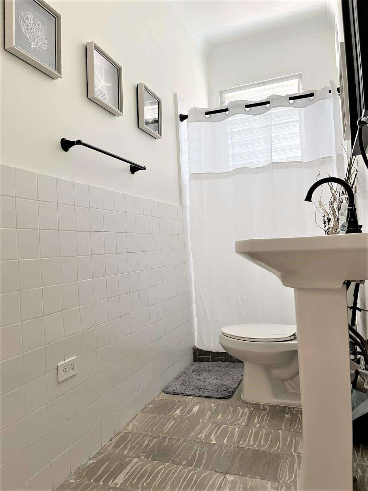 A bathroom with white tiled walls, a toilet, a sink, and framed pictures on the wall.