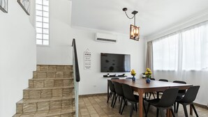 Een eethoek met een houten tafel en zwarte stoelen, een aan de muur bevestigde televisie en een trap naar een hoger gelegen verdieping.