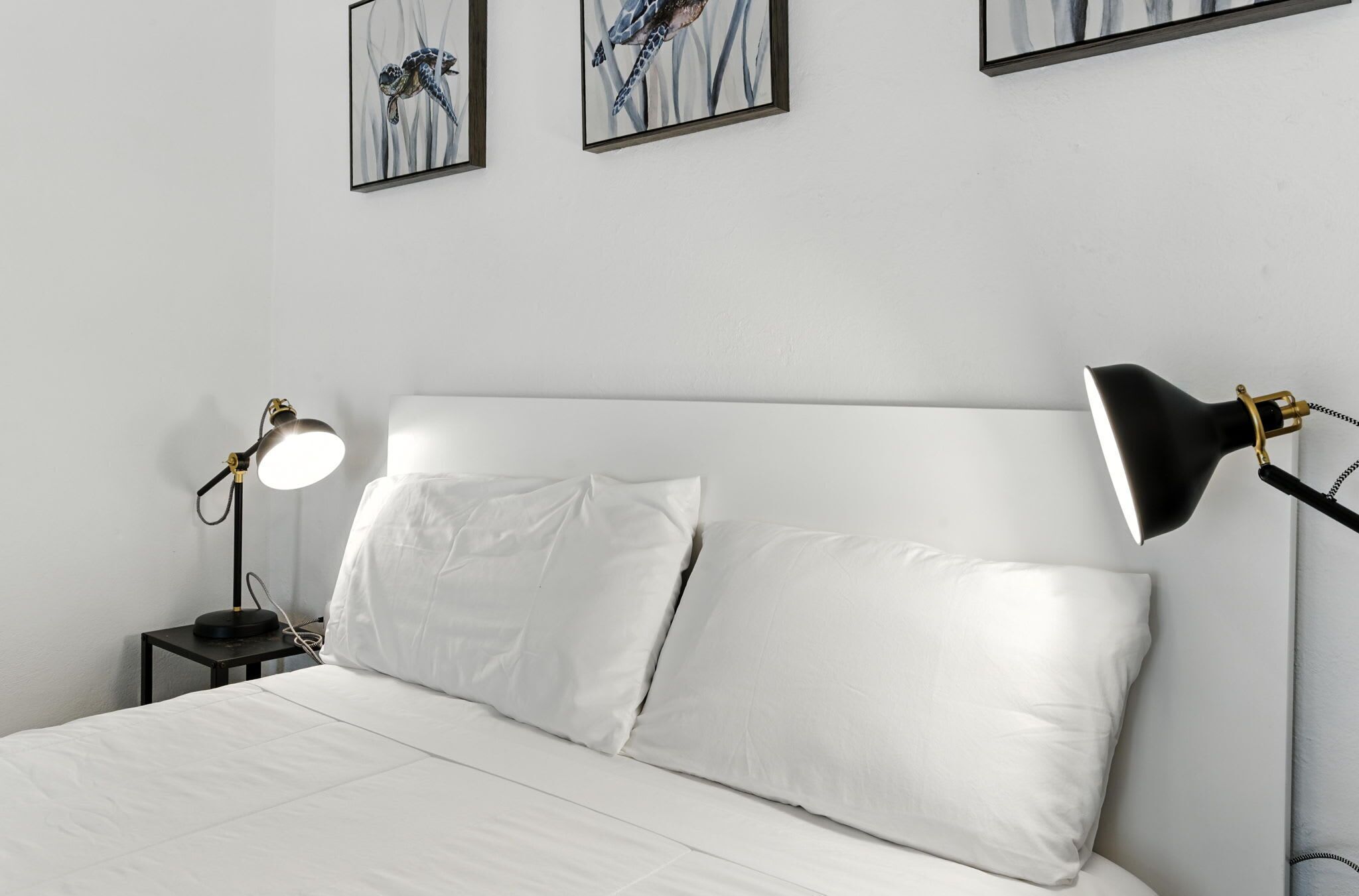 A bed with white linens, two bedside lamps, and two framed artworks on the wall.
