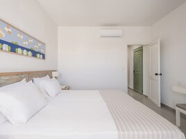 Habitación de hotel con cama, mesita de noche, un cuadro, equipo de aire acondicionado y puerta que da a otra habitación.