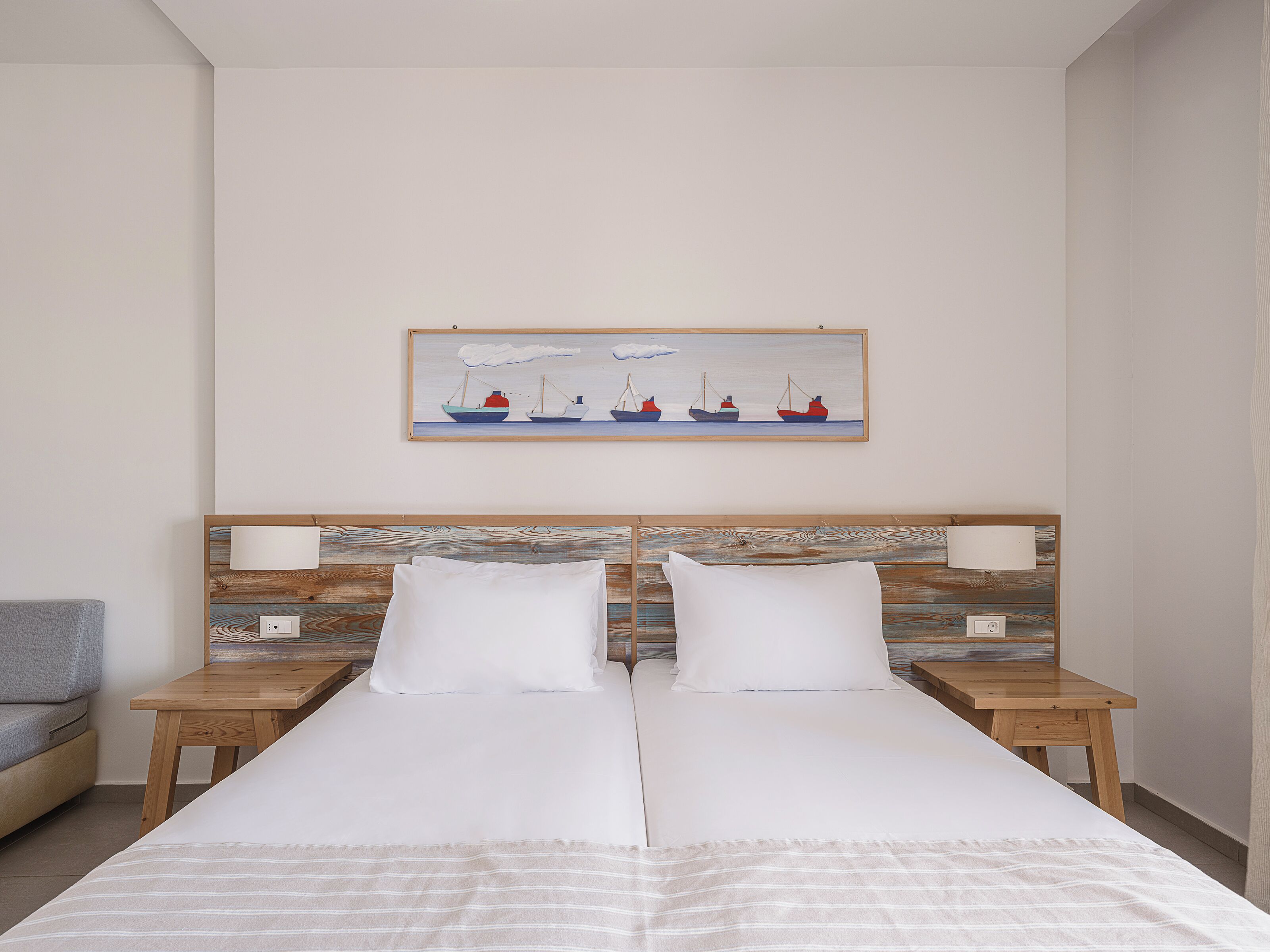 A double bed with white linens, two bedside tables with lamps, a painting of boats on the wall, and a grey armchair.