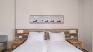 A double bed with white linens, two bedside tables with lamps, a painting of boats on the wall, and a grey armchair.