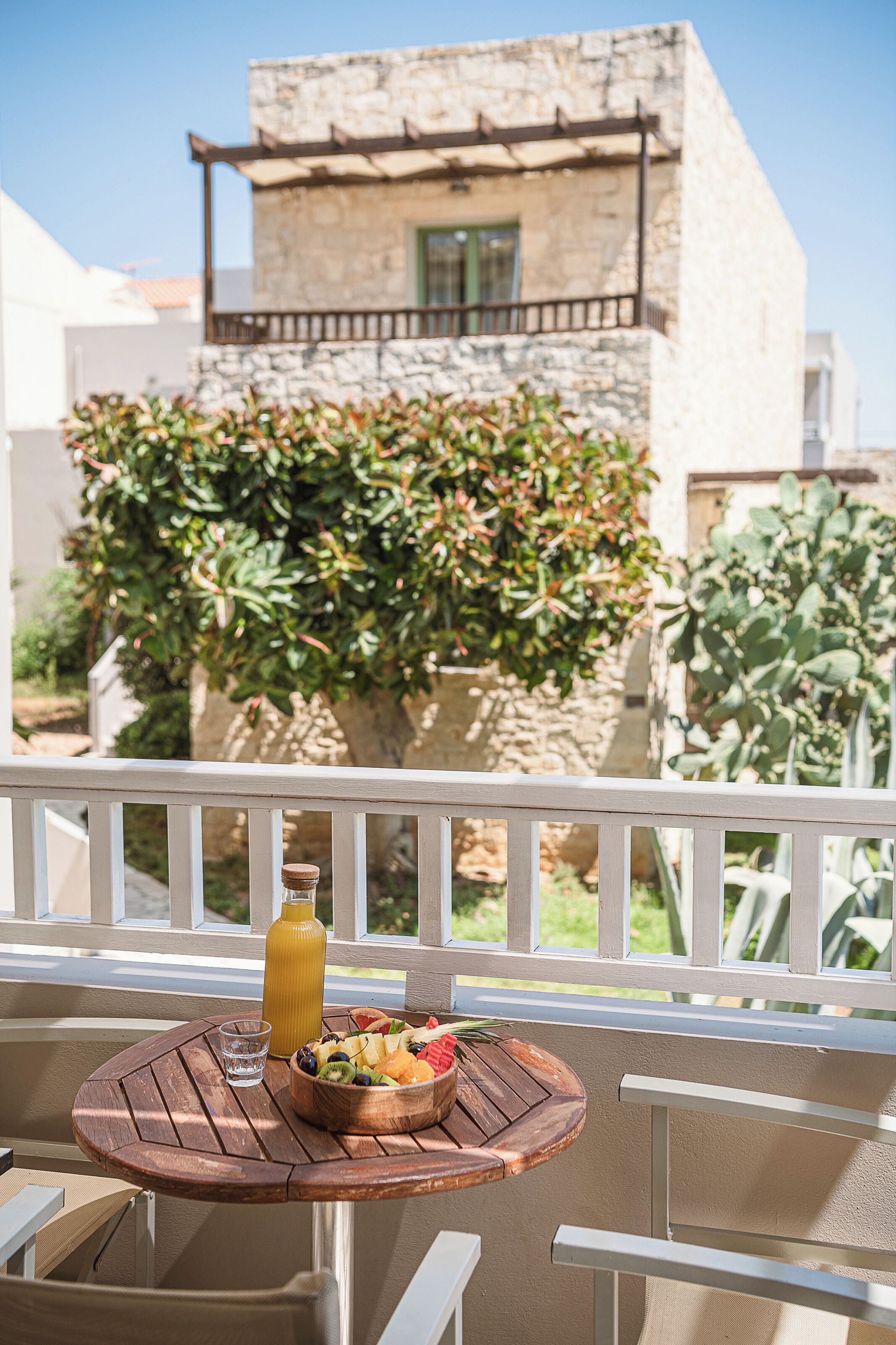 Une table avec un panier de fruits et une bouteille de jus sur un balcon donnant sur un bâtiment en pierre et sur de la verdure.