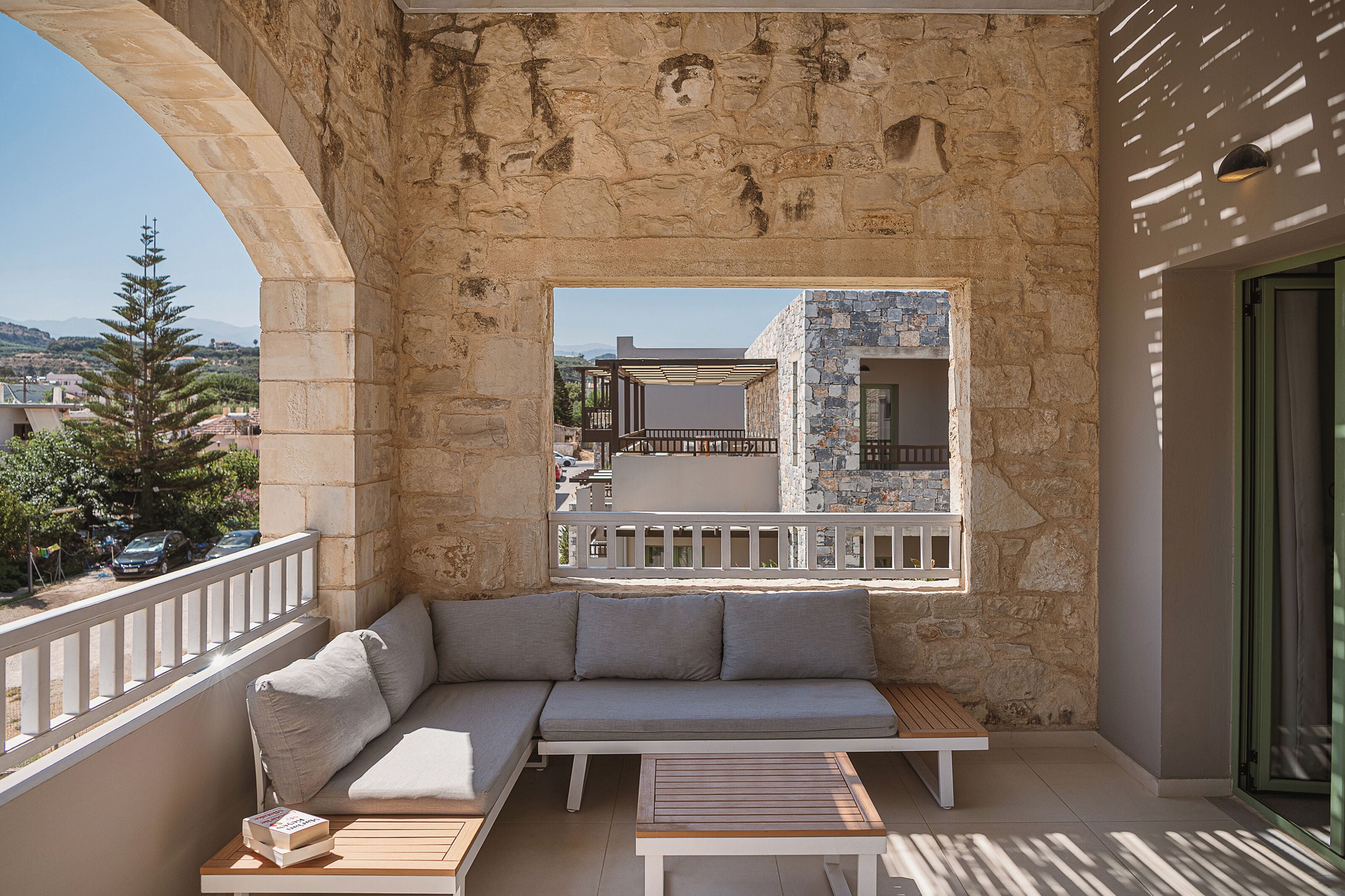 Un balcón con un muro de piedra, una zona de estar con cojines y vistas a un edificio con balcón.