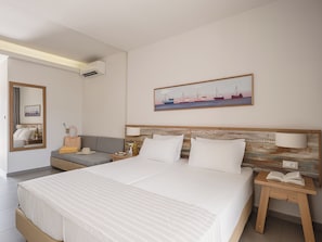A hotel room with a large bed, a bedside table with a lamp, a sofa, a small table with a hat and a bottle, and a painting on the wall. - Erato Beach Hotel Adults Only (Platanias)