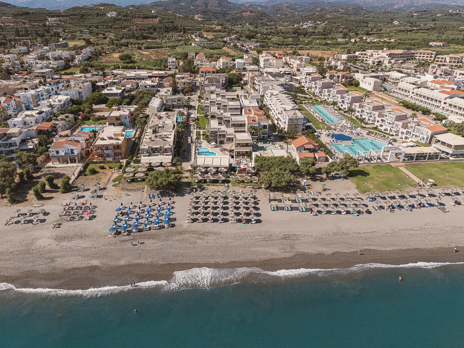 An aerial view of a coastal resort with multiple buildings, a beach with umbrellas and sun loungers, and a swimming pool.