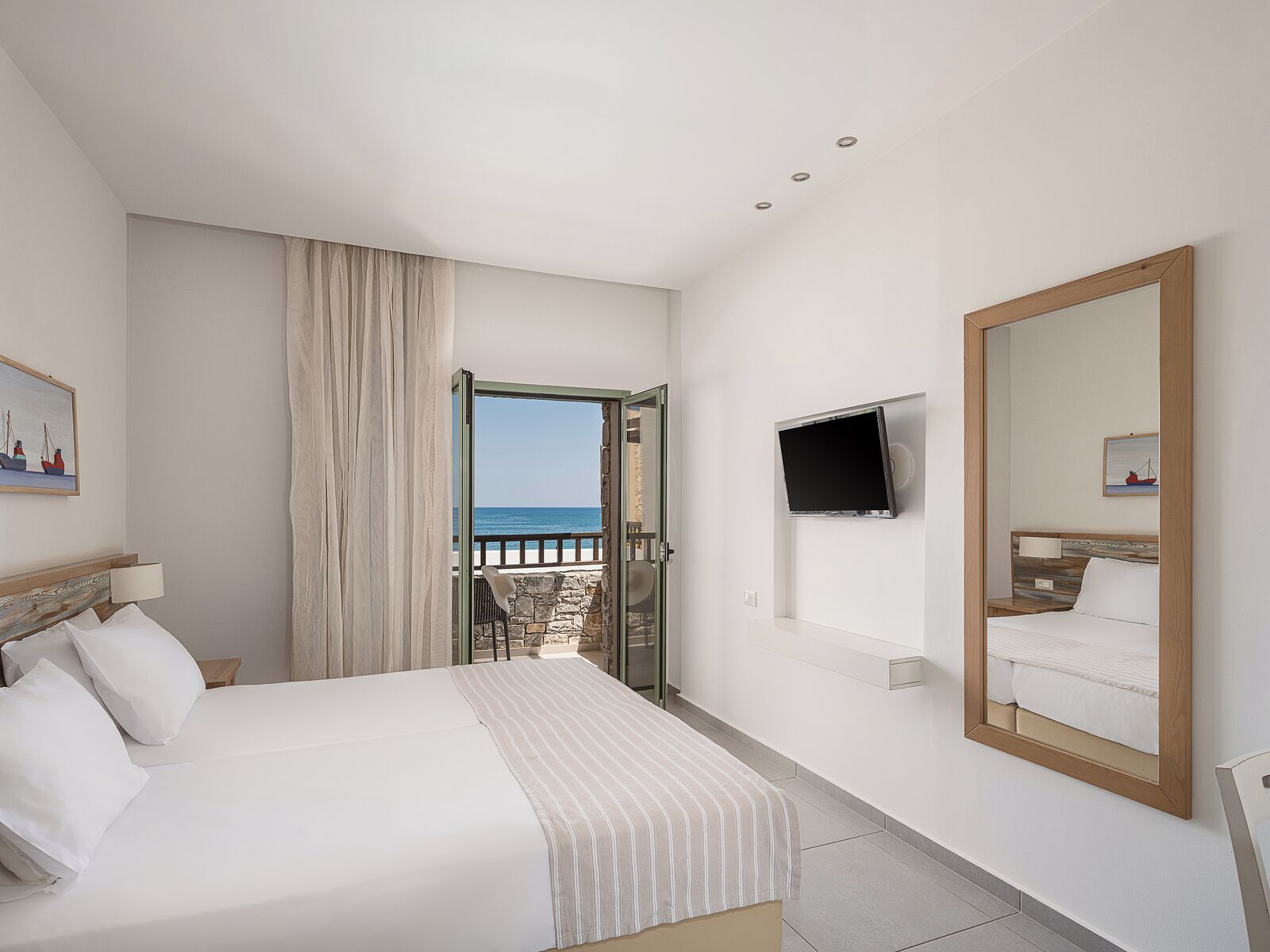 Habitación de hotel con cama, televisor, un espejo grande y vista al mar a través de una puerta abierta.