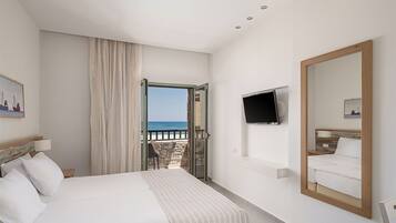 Habitación de hotel con cama, televisor, un espejo grande y vista al mar a través de una puerta abierta.
