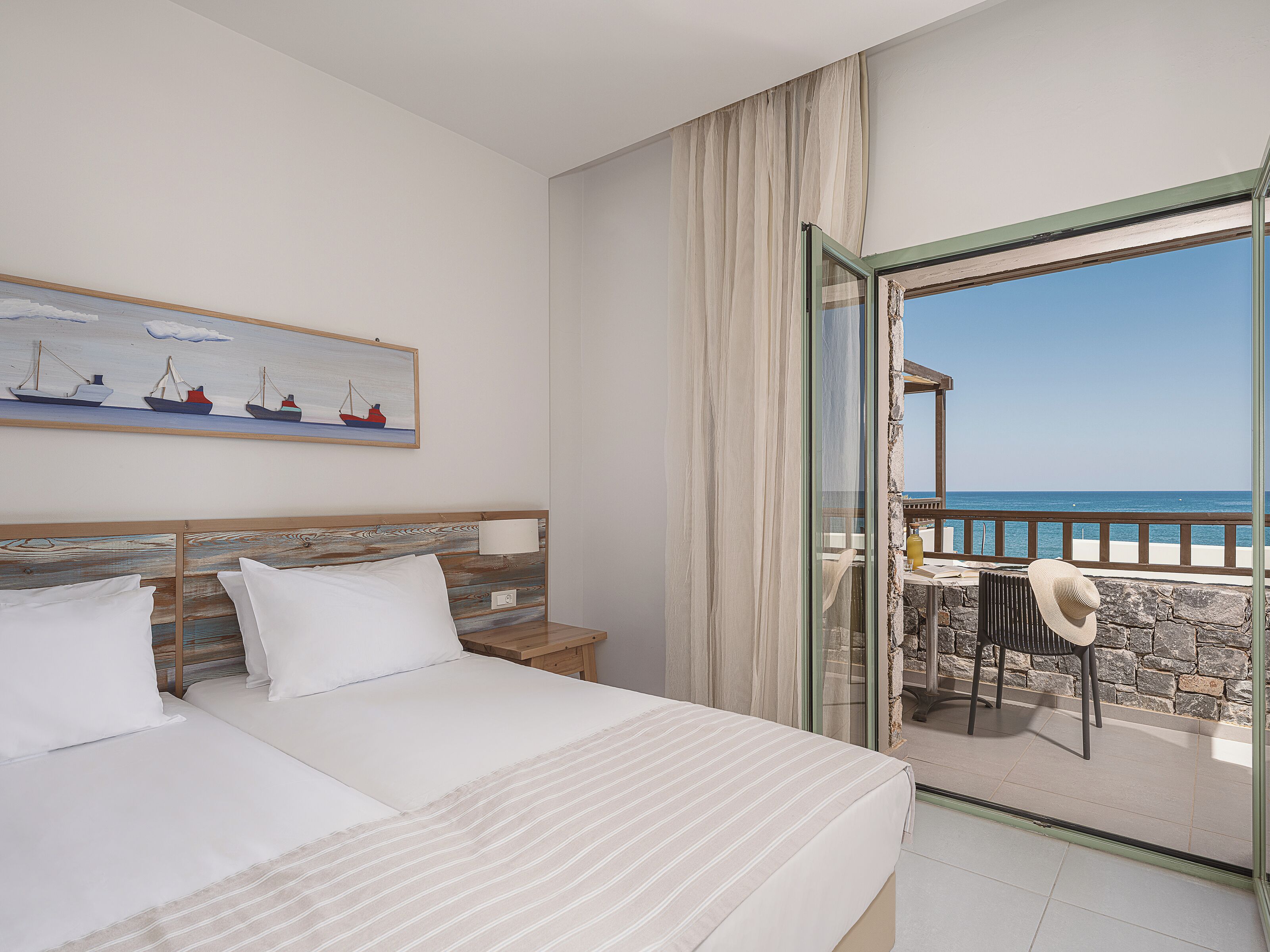 Una habitación de hotel con una cama, un cuadro de barcos, un balcón con vistas al mar y una mesa con una silla.