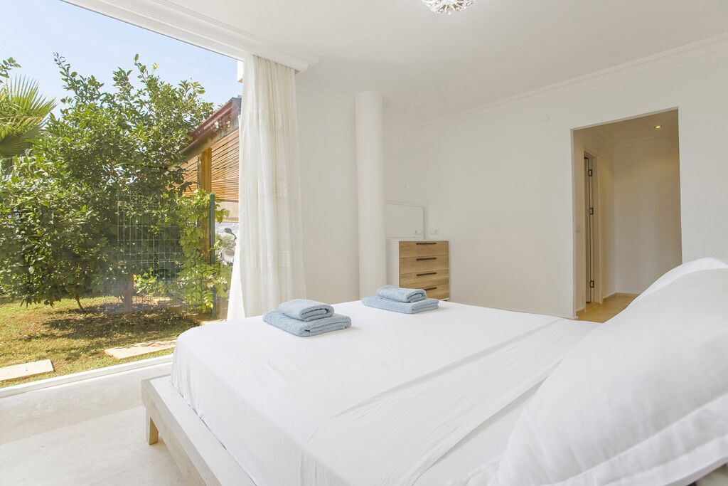 Un dormitorio con un ventanal grande, una cama con ropa de cama blanca, una mesita de noche de madera y vistas a un jardín.