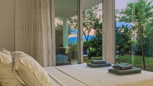 Une chambre avec une grande fenêtre, un lit avec des oreillers et une vue sur la verdure et un bâtiment à l’extérieur.