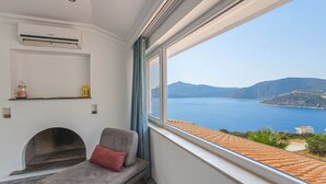 Une pièce avec vue sur un plan d’eau et des montagnes, une cheminée et un canapé agrémenté d’un coussin rouge.