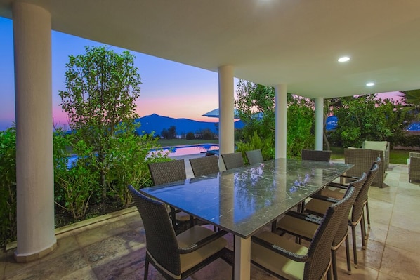 An outdoor patio with a glass-top table and wicker chairs, surrounded by greenery and overlooking a scenic view of mountains and a body of water.