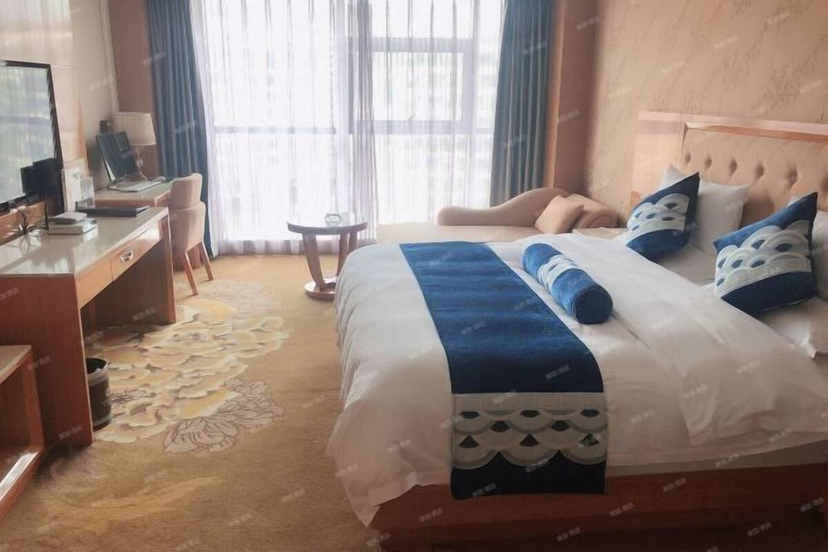 A hotel room with a bed, a desk, a chair, a small table, and a large window with curtains.