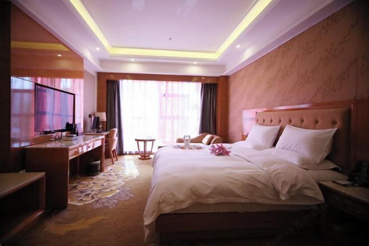 Quarto de hotel com cama, mesa de cabeceira, escrivaninha, cadeira e televisão.