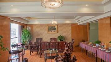 A hotel dining area with wooden flooring, a chandelier, a long table with a striped tablecloth, and a large potted plant.