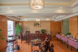 A hotel dining area with wooden flooring, a chandelier, a long table with a striped tablecloth, and a large potted plant. - Shuangzixing Hotel (Mojiang)